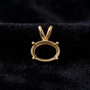 May include: A gold pendant setting featuring an oval-shaped frame with four prongs designed to hold a gemstone. The piece includes a V-shaped bail at the top for attachment to a chain, showcasing a polished metallic finish throughout the structure.