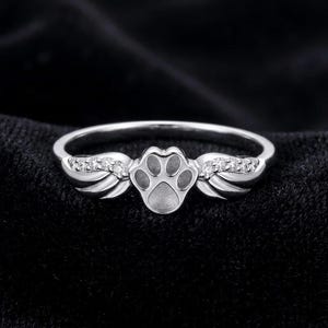 May include: A silver-toned ring featuring a central paw print design flanked by two stylised wings. The wings are embellished with small, clear, sparkling stones set along the upper edges, creating a detailed and symmetrical metallic band.