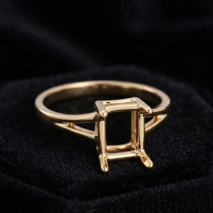 May include: A gold ring setting featuring a rectangular prong head designed to hold a gemstone. The band is smooth and polished, with a split shank design that connects to the four-prong setting, creating an open structure for a stone.