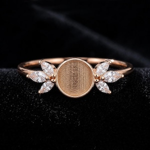 Op de afbeelding: Een ros&eacute;gouden ring met een centrale ronde zetting met een textuurrijk, verzonken oppervlak. De ring is aan beide zijden versierd met een cluster van drie heldere, marquise-geslepen stenen die in een bladvorm langs de band zijn geplaatst.