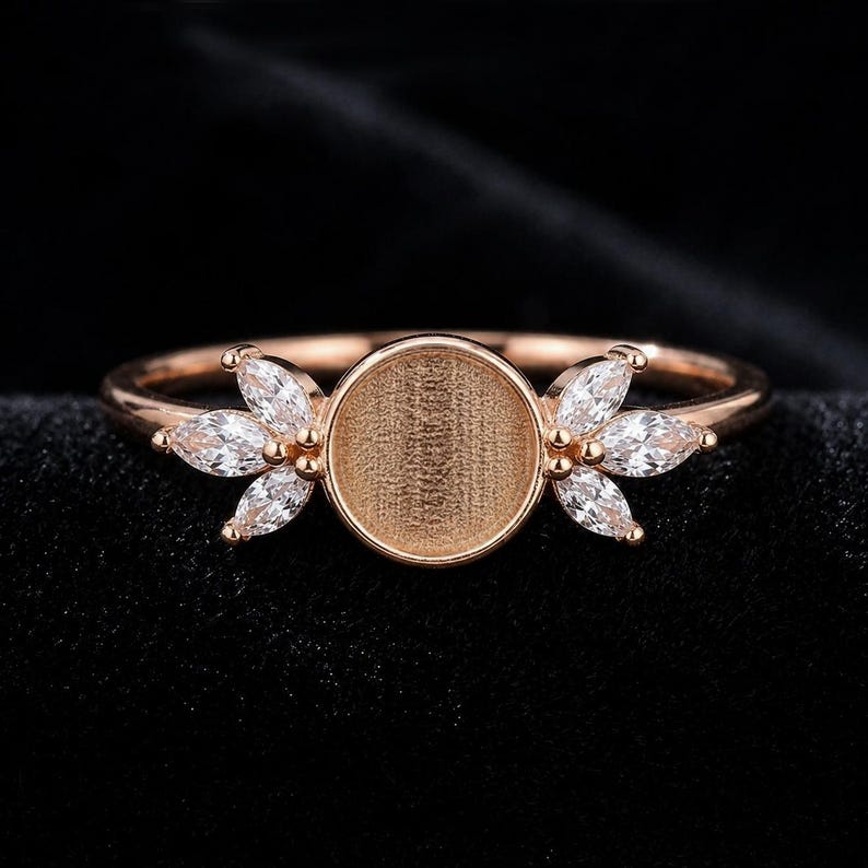 Op de afbeelding: Een ros&eacute;gouden ring met een centrale ronde zetting met een textuurrijk, verzonken oppervlak. De ring is aan beide zijden versierd met een cluster van drie heldere, marquise-geslepen stenen die in een bladvorm langs de band zijn geplaatst.