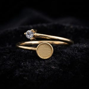 Peut inclure: Une bague ouverte en or avec une pierre précieuse ronde et transparente sertie sur une extrémité et un disque plat circulaire en or sur l'autre. La bague présente une finition dorée polie et une conception ouverte permettant un ajustement.