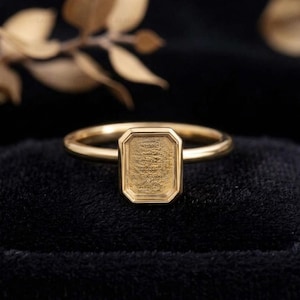 May include: A gold ring featuring a rectangular bezel setting with a textured, recessed center. The band is smooth and polished, connecting to the octagonal-shaped head. The metal has a warm, metallic yellow tone throughout the entire piece.