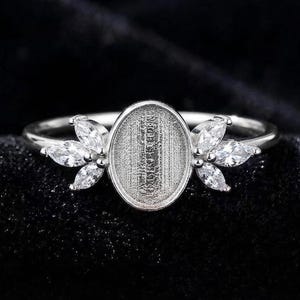 Op de afbeelding: Een zilverkleurige ring met een centrale ovale zetting met een getextureerde basis, geflankeerd aan weerszijden door drie heldere stenen in marquise-slijpvorm, gerangschikt in een bladvormig patroon. De ringband is dun en gepolijst.