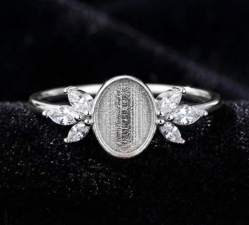 Op de afbeelding: Een zilverkleurige ring met een centrale ovale zetting met een getextureerde basis, geflankeerd aan weerszijden door drie heldere stenen in marquise-slijpvorm, gerangschikt in een bladvormig patroon. De ringband is dun en gepolijst.