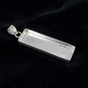 May include: A rectangular silver pendant tray featuring a smooth, flat base with raised edges and a small attached bail at the top. The metal surface has a polished, reflective finish, designed to hold an inlay or cabochon within its frame.