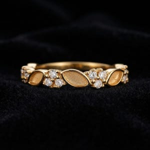 May include: A gold band ring featuring an alternating pattern of marquise-shaped textured gold segments and clusters of three small round clear stones. The design repeats around the circumference of the band, creating a continuous decorative motif.