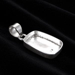 Puede incluir: Un colgante de metal de color plateado que presenta un engaste rectangular hueco con un acabado liso y pulido. El colgante incluye una anilla redondeada integrada en la parte superior, diseñada para sujetarse a una cadena o cordón.