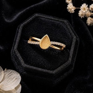 May include: A gold ring featuring a teardrop-shaped setting with a textured, matte finish at the centre. The band is split into two delicate, crossing arms, with one arm encrusted with small, clear, sparkling stones along its length.
