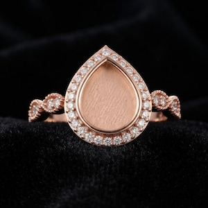 May include: A rose gold ring featuring a pear-shaped setting with a textured, flat centre. The pear-shaped frame is surrounded by a halo of small, clear, round-cut stones. The band has a decorative, twisted design adorned with additional small stones.