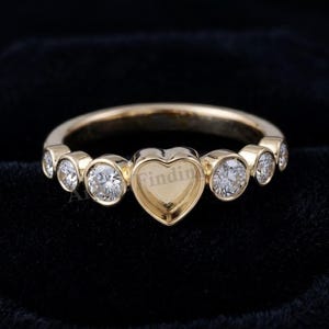 May include: A gold ring featuring a central heart-shaped setting flanked by three round bezel-set stones on each side. The band is polished gold, and the setting is designed to hold a gemstone. The word Finding is visible on the heart setting.
