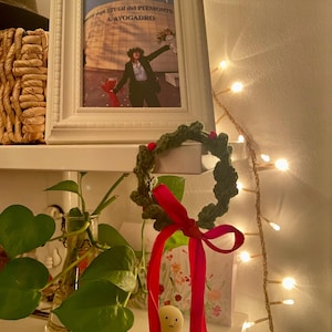 May include: A white framed photograph with text in Italian, a small green wreath with red berries and a red ribbon, and a small figurine with green pants and a yellow body. A string of lights is in the background.