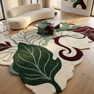 Könnte beinhalten: Ein Teppich mit unregelmäßiger, organischer Form und einem großflächigen botanischen Muster. Das Design zeigt dunkelgrüne, salbeigrüne und dunkelburgunderrote Blattmotive auf cremefarbenem Grund mit geschnitzten, strukturierten Details.