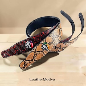 May include: Two guitar straps with a snakeskin pattern. One strap is red and black, the other is brown and tan. Each strap has a black backing and a decorative patch with the text "Rusty Tone". The straps are on a wooden surface.