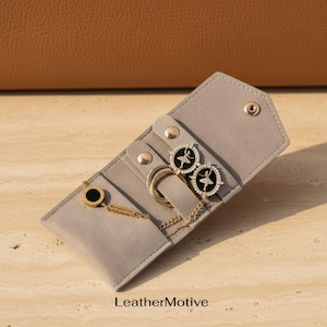 May include: A light gray suede jewelry organizer case with a snap closure containing gold-toned jewelry, including two circular ballerina-themed pendants with black centers and a gold chain bracelet with a black circular charm. Text reads LeatherMotive.