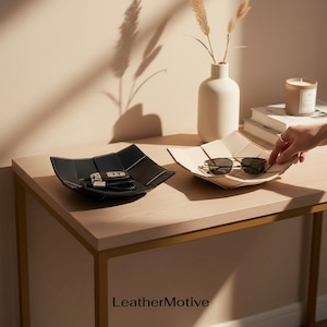 May include: Two square valet trays, one black and one beige, featuring a folded geometric design with visible stitching. The black tray holds a set of car keys, while the beige tray contains a pair of dark-rimmed sunglasses. The text LeatherMotive is visible.