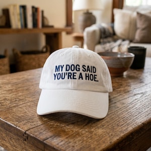 Può includere: Cappellino da baseball bianco con visiera curva. Il cappellino presenta la scritta "MY DOG SAID YOU'RE A HOE" in blu navy. Il cappellino è appoggiato su un tavolo di legno.