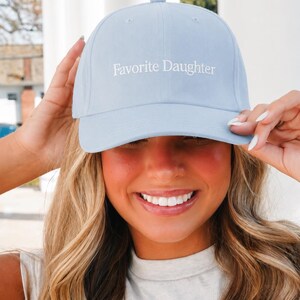 May include: A light blue baseball cap featuring the text Favorite Daughter embroidered in white thread on the front panel. The cap has a structured crown, a curved brim, and a button on top, providing a clean and simple design for casual wear.