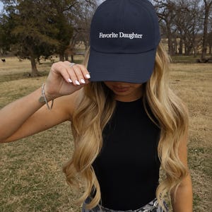 May include: A navy blue baseball cap featuring the white embroidered text Favorite Daughter on the front panel. The cap has a structured crown, a curved brim, and a button on top, providing a classic athletic look with clean white lettering.