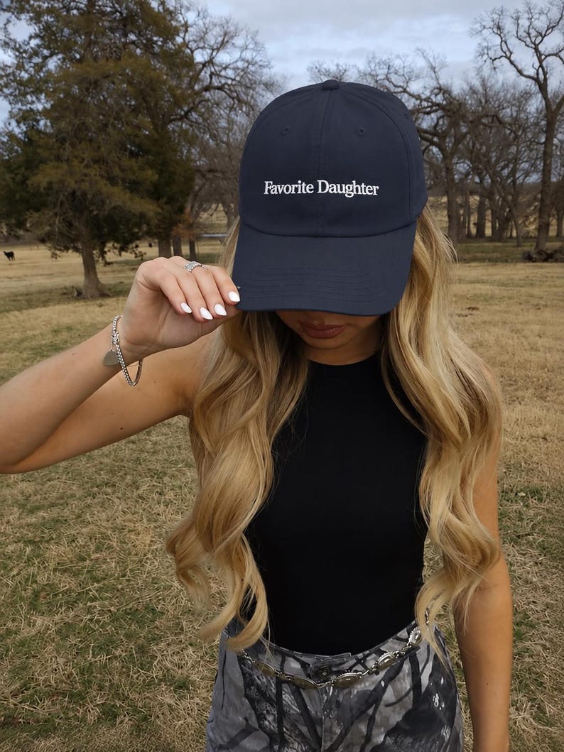 May include: A navy blue baseball cap featuring the white embroidered text Favorite Daughter on the front panel. The cap has a structured crown, a curved brim, and a button on top, providing a classic athletic look with clean white lettering.