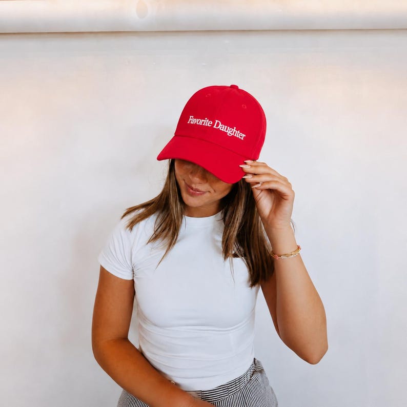 May include: A bright red baseball cap featuring the text Favorite Daughter embroidered in white lettering across the front panel. The cap has a structured crown, a curved brim, and a matching red button at the top center of the headwear.