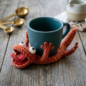 Könnte beinhalten: Eine türkisfarbene Keramiktasse steht auf einem handgefertigten, orangefarbenen Häkellizard-Untersetzer mit weißen und schwarzen Augen und einem roten Maul. Der Untersetzer befindet sich auf einer rustikalen Holzoberfläche.