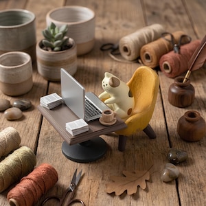 May include: A miniature figurine of a cream-colored cat with brown spots sitting in a yellow armchair at a small brown desk. The desk holds a silver laptop, a tiny cup of coffee, and two stacks of white paper. Nearby are spools of yarn and scissors.