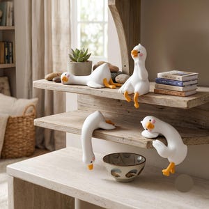 Peut inclure: Quatre figurines d'oies blanches avec des becs et des pattes orange dans des poses variées. L'une est allongée, une autre assise, une troisième pend la tête en bas d'une étagère et la dernière s'accroche au bord. Elles sont sur des étagères.