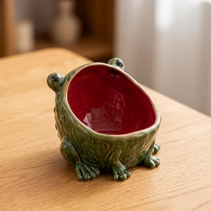 May include: A ceramic bowl shaped like a frog with a textured green exterior and a smooth, deep red interior. The frog features two prominent eyes on the top rim and molded front legs resting at the base, creating a wide, open-mouthed vessel.