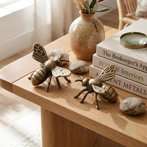 May include: Two antique brass bee figurines with detailed wings and segmented bodies. The bees are positioned on a light wood surface alongside a ceramic vase, smooth stones, and a stack of three books titled The Beekeeper's Apprentice, Bespoke Interiors, and Ancient Metals.