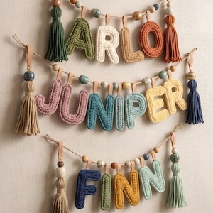 May include: Three decorative garlands featuring crocheted letters spelling ARLO, JUNIPER, and FINN. Each garland is strung on twine with wooden beads and finished with large yarn tassels at both ends. The letters are in various colors including green, white, tan, orange, pink, blue, yellow, and light green.