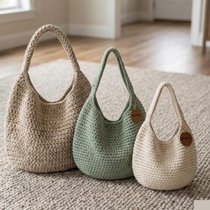 Op de afbeelding: Drie gehaakte draagtassen in verschillende maten en kleuren. De grootste is beige, de middelste saliegroen en de kleinste gebroken wit. Elke tas heeft een afgeronde vorm, een gevlochten handvat en een klein, rond label met "ORIGINAL".