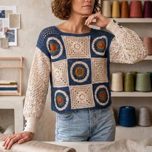 May include: A handmade crochet sweater featuring a patchwork design. The sweater has a square neckline and long sleeves. The body of the sweater is made of blue and cream-colored squares with circular and floral patterns. The sleeves are cream-colored with button details.