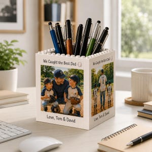 May include: White block-style pen holder with a baseball theme. The holder features photos of a father and sons, with the text "We Caught the Best Dad" and "Love, Tom & David". Assorted pens are inside the holder.
