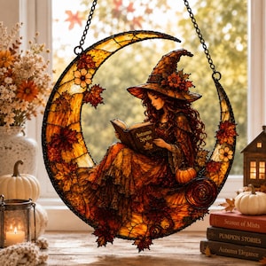 May include: A stained glass suncatcher shaped like a crescent moon featuring a witch with long hair wearing a pointed hat and reading a book titled Autumn Spells. The piece is decorated with orange and red maple leaves and a small pumpkin.