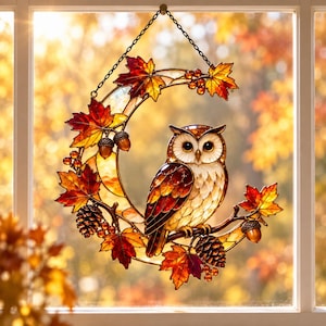 May include: A stained glass suncatcher shaped like a crescent moon, featuring a perched owl surrounded by autumn maple leaves, pinecones, and acorns in shades of orange, red, and brown. It hangs from a black metal chain against a window.
