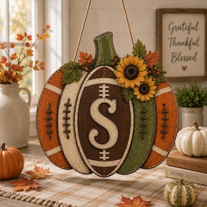 May include: A hanging pumpkin-shaped wall sign featuring a football design with a central letter S. The sign is decorated with sunflowers and green leaves, colored in shades of orange, cream, brown, and green with a distressed finish.