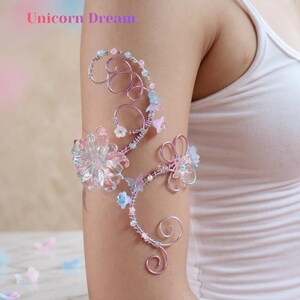 May include: A delicate arm cuff with a floral design. The cuff is made of silver wire and features pink, blue, and clear flowers and beads. The text "Unicorn Dream" is displayed in the upper left corner. The cuff is worn on an arm.