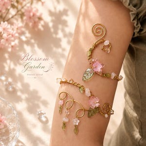 May include: A gold-toned arm cuff with floral and leaf accents. The cuff features pink flower and green leaf details, along with clear and pink crystal beads. The text "Blossom Garden PHONE STRAP" is visible in the image.