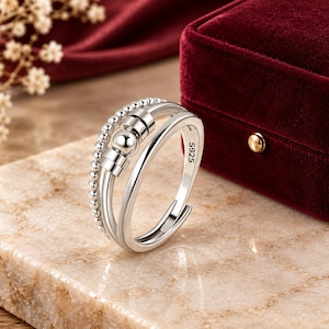 May include: A silver-colored open ring featuring a double-band design. One band consists of a row of small beads, while the other is a smooth, polished band. Three movable silver beads are positioned on the bands. The interior is stamped S925.
