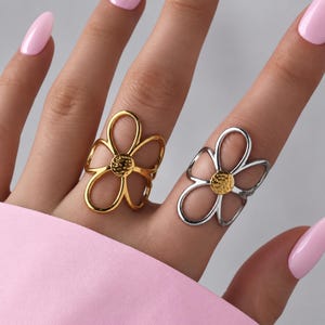 Pode incluir: Dois anéis em forma de flores vazadas, um em tom dourado polido e outro em tom prateado. Cada anel apresenta cinco pétalas arredondadas e alongadas, além de uma peça central circular texturizada que combina com o tom dourado do primeiro.