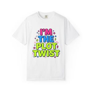 Puede incluir: Camiseta blanca con la frase "I'M THE PLOT TWIST" en letras de burbujas rosas, azules y verdes. El texto está decorado con estrellas y rayos. La camiseta tiene cuello redondo y mangas cortas.