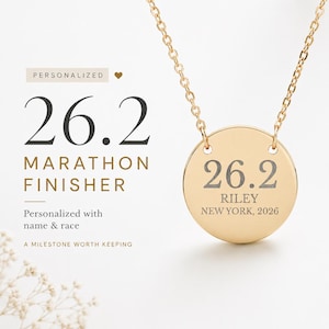 May include: Gold-coloured necklace with a round pendant. The pendant features the number "26.2" and the text "RILEY NEW YORK, 2026". The necklace is designed for marathon finishers. The text "MARATHON FINISHER" is also visible.