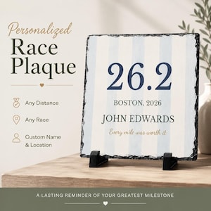 Puede incluir: Una placa de pizarra cuadrada con bordes naturales y rugosos, expuesta sobre dos soportes de plástico negro. La superficie presenta rayas verticales azul claro y blancas, el texto 26.2, BOSTON, 2026, JOHN EDWARDS y la frase Every mile was worth it.