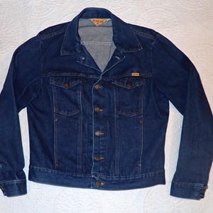 May include: A dark blue denim jacket with a button-up front, two chest pockets, and two side pockets. The jacket has a classic collar and long sleeves. The brand name is visible on a small tag on the chest pocket.