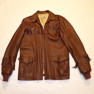 May include: A brown leather jacket with fringe detailing on the chest, sleeves, and cuffs. The jacket has a zip-up front, two front pockets, and a collared neckline. The cuffs are a darker brown color.