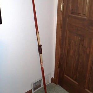 May include: A red wooden bow with a white stripe down the center and a brown leather grip. The bow is leaning against a wall.