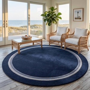 May include: A large, circular area rug featuring a deep navy blue center surrounded by a wide, light gray border. The rug has a solid, uniform texture and is positioned on a light-colored wooden floor in a room with rattan furniture.