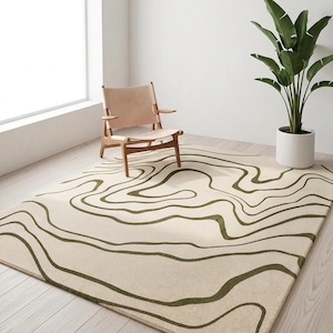 May include: A rectangular area rug featuring a cream-coloured base with dark olive green wavy, organic lines that traverse the surface in a topographic pattern. The rug is positioned on a light-coloured wooden floor in a bright, open room.