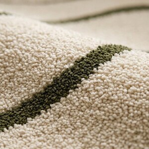 May include: A close-up view of a textured rug featuring a thick, cream-coloured pile with distinct, curved stripes in a deep olive green. The surface shows a dense, tufted construction with a soft, looped appearance throughout the fabric.